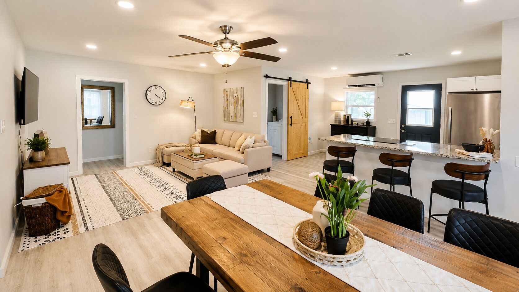 A modern living room with a sofa, dining table, and chairs, a kitchen with a refrigerator, and a ceiling fan.