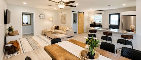 A modern living room with a sofa, dining table, and chairs, a kitchen with a refrigerator, and a ceiling fan.