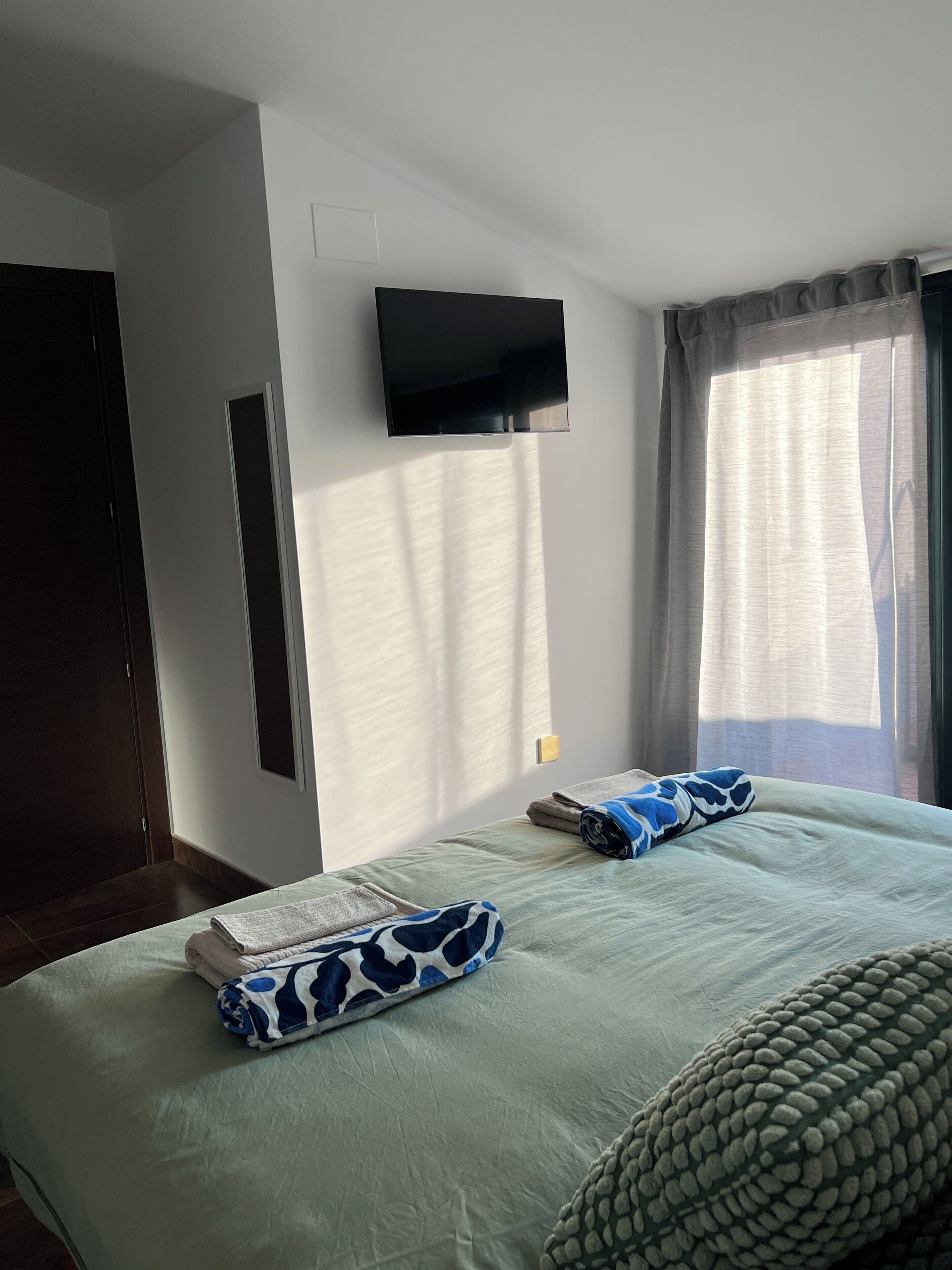 A hotel room with a bed, a flat-screen TV mounted on the wall, a window with sheer curtains, and a door.