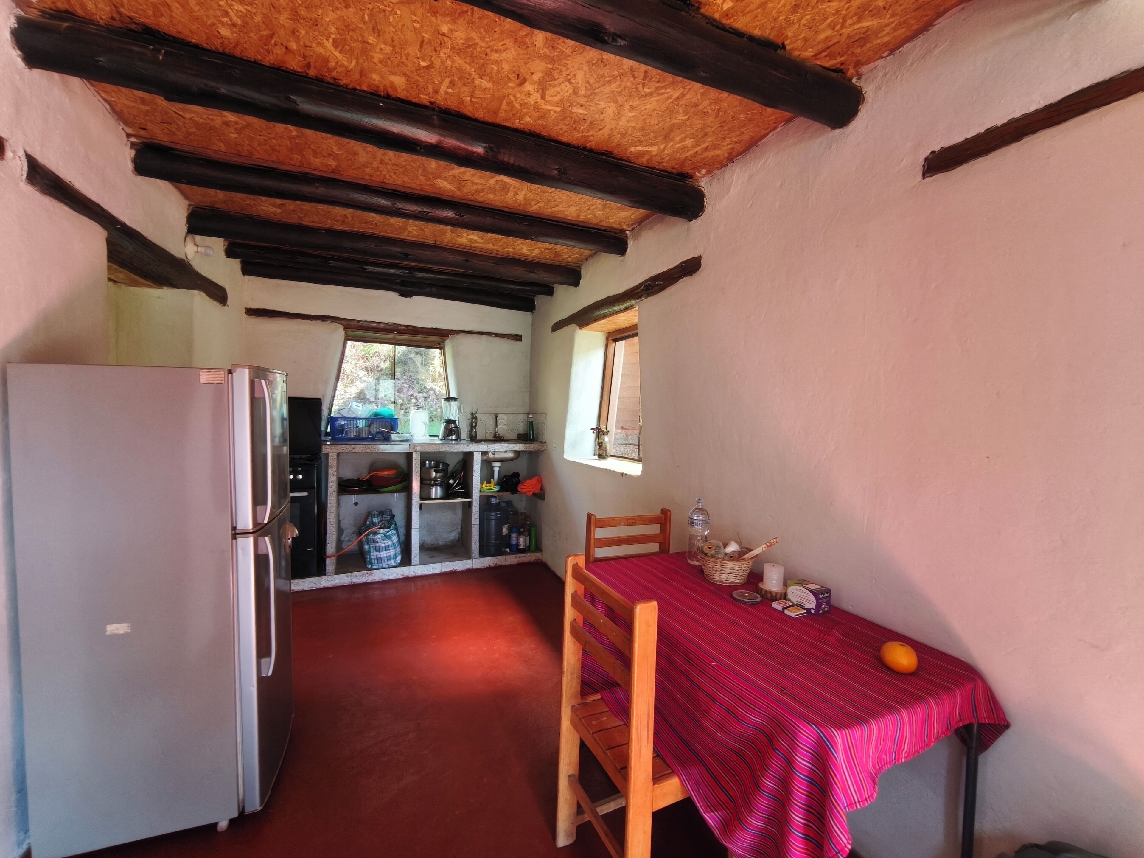 A compact kitchen with a refrigerator, wooden beams, a dining table with a red tablecloth, and a window.