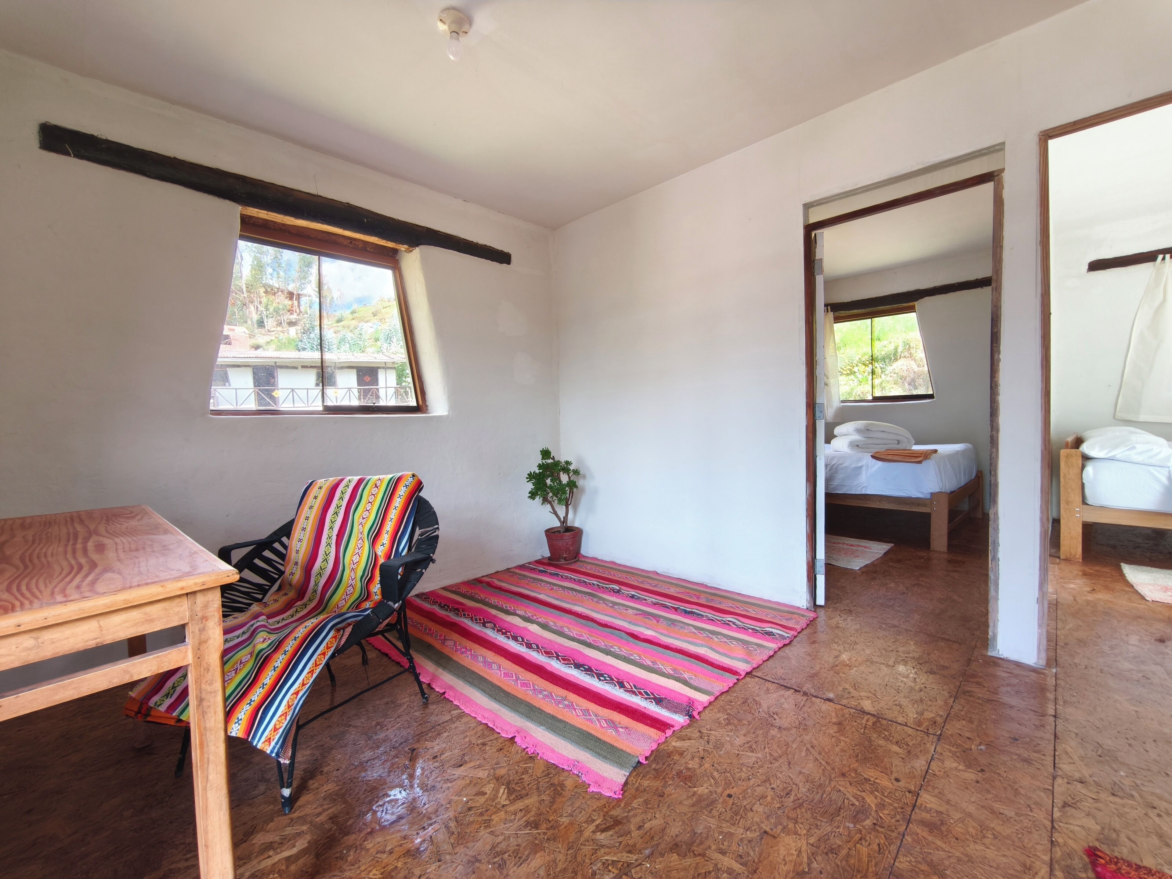A room with a wooden table, a colorful striped chair, a potted plant, a bed with white bedding, and a view through an open door.