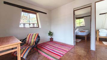 A room with a wooden table, a colorful striped chair, a potted plant, a bed with white bedding, and a view through an open door.