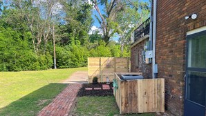 A brick pathway leading to a wooden fence, a small garden area with a planter, and a brick building with a window and door.