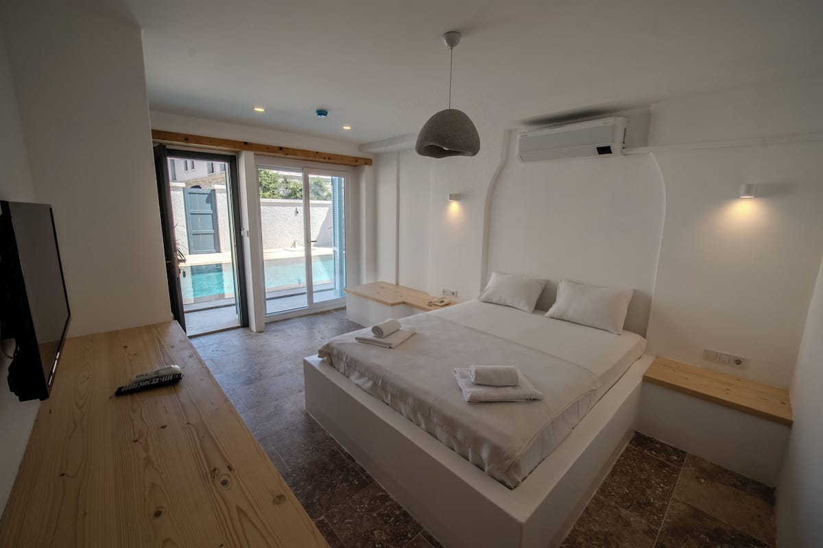Un dormitorio con una cama grande, una mesita de noche de madera, un control remoto y un ventanal corredero que da a un balcón.