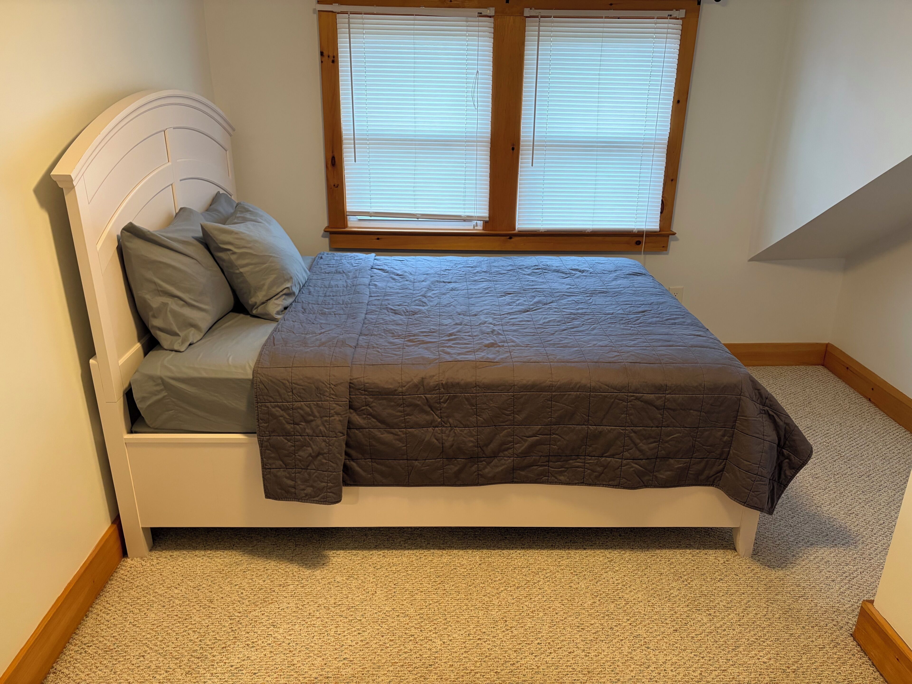 A single bed with a headboard, a window with blinds, and a carpeted floor.