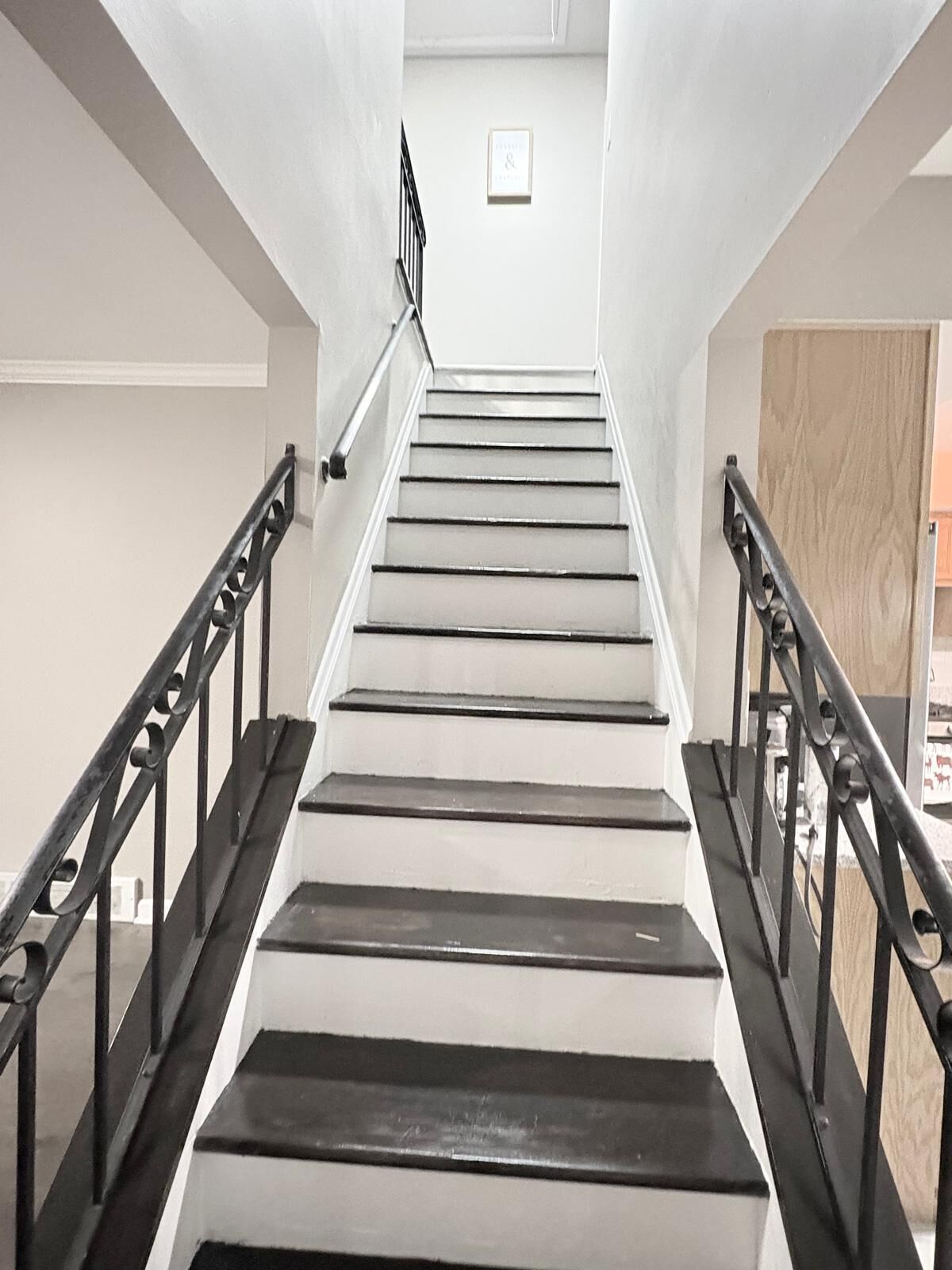 Un escalier avec des rampes noires et des marches blanches, menant à un palier où est accrochée une image encadrée.