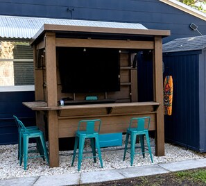 Eine hölzerne Bar im Freien mit Fernseher, zwei türkisfarbenen Stühlen und einer Tiki-Masken-Dekoration.