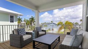 A covered outdoor seating area with wicker furniture, a table, and a view of palm trees and a house. - Freshly Rebuilt Beach Escape — 4 Bedrooms, Pool & Island Vibes (Fort Myers Beach)