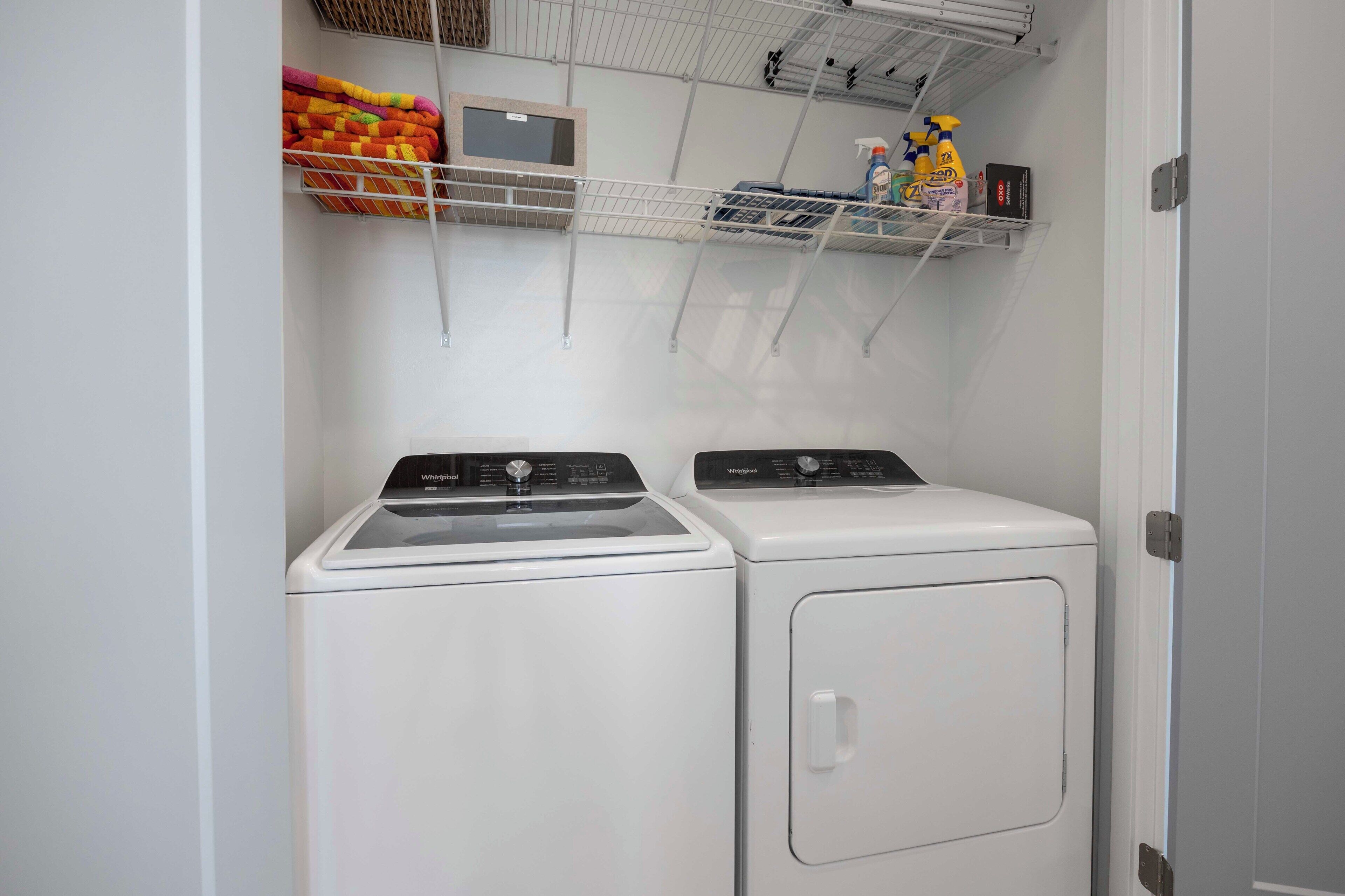 A laundry room with two front-loading washers and dryers, a shelf with cleaning supplies, and a basket of folded blankets.