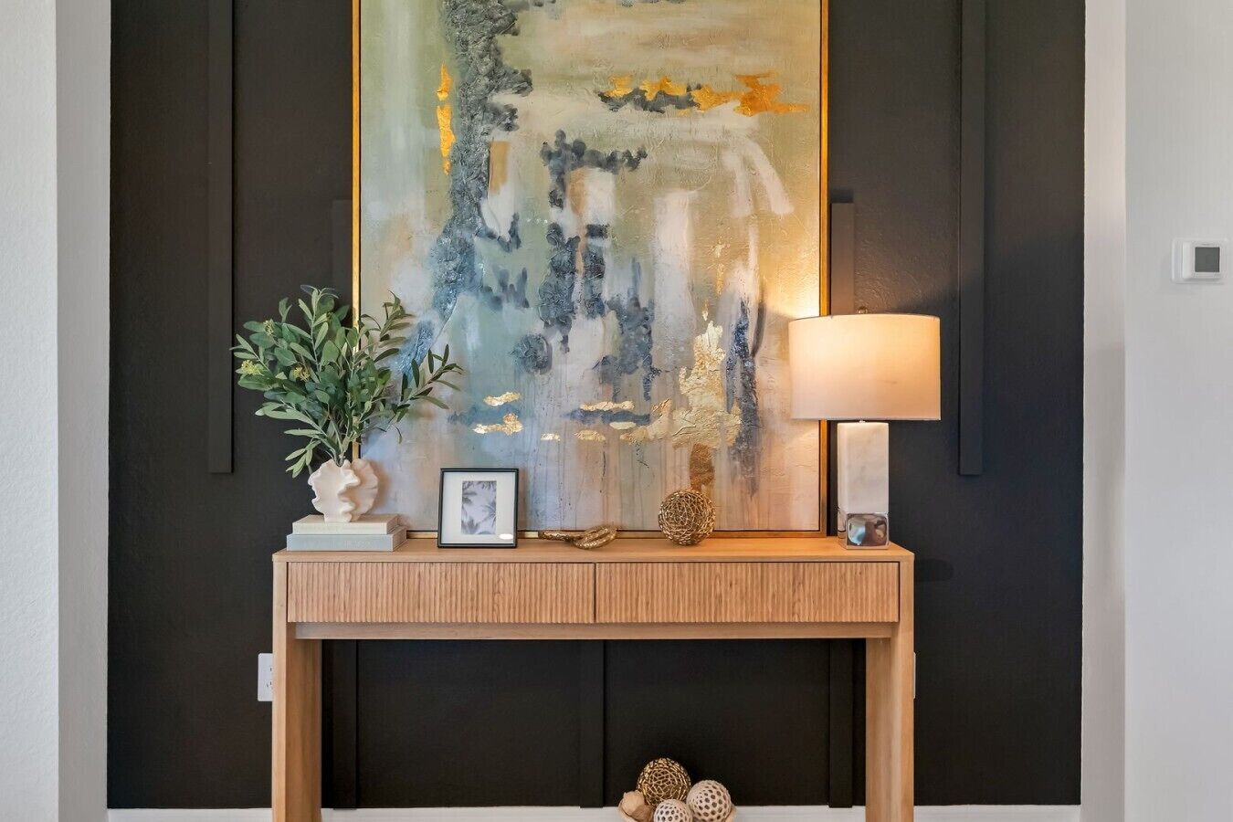 Une console en bois ornée d’un tableau encadré, d’une plante en pot, d’objets décoratifs et d’une lampe fixée au mur.