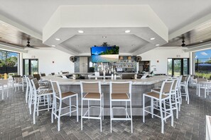 A bar area with white bar stools, a large TV screen, and a kitchen in the background. - Luxe Golf Coach Home Near Venice Beach FL (Venice FL)