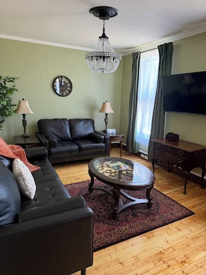 Ein Wohnzimmer mit einem schwarzen Ledersofa, einem Glastisch, einer Chandelüster, einem Fernseher und einer Wanduhr.