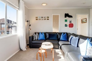 A living room with a black leather sofa, a white coffee table, and a small wooden chair. There are wall-mounted pictures and a door with a red decoration. - St Heliers 2BR Escape with Views & Parking (Auckland Central Suburbs)