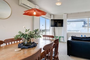 A dining area with a wooden table, chairs, a vase with flowers, a red pendant light, and a TV mounted on the wall. - St Heliers 2BR Escape with Views & Parking (Auckland Central Suburbs)