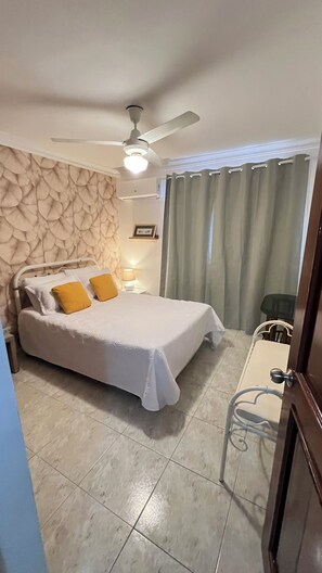 Quarto com cama, escrivaninha, cadeira, ventilador de teto e janela com cortinas.