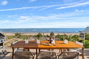 Un tavolo di legno con sedie in vimini, un barbecue e bottiglie su un patio con vista sull'oceano.