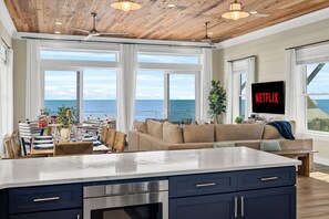 Ein modernes Wohnzimmer mit großem Fenster mit Meerblick, einem Küchenbereich und einem Flachbildfernseher, der 'NETFLIX' anzeigt.