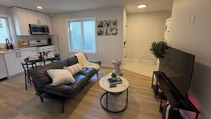 Sala de estar moderna com sofá de couro, mesa de centro, televisão e área de cozinha com armários e eletrodomésticos brancos.