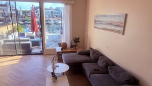 Ein modernes Wohnzimmer mit einem grauen Sofa, einem kleinen weißen Tisch mit einer Pflanze und Blick auf eine Marina durch große Glasüren.