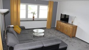 Een woonkamer met een grijze bank, een klein wit tafeltje, een televisie op een houten kast en een raam met gordijnen.