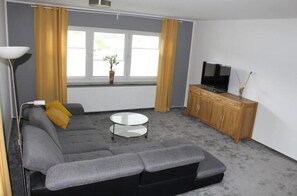 Ein Wohnzimmer mit grauem Sofa, einem kleinen weißen Tisch, einem Fernseher auf einer Holzkommode und einem Fenster mit Vorhängen.