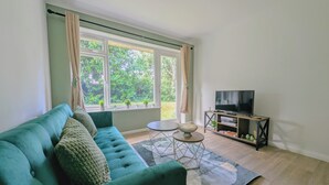 Un salon avec un canapé de couleur turquoise, un téléviseur posé sur un meuble en bois, une petite table et une fenêtre avec des rideaux.