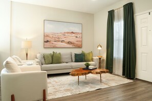 Una sala de estar con sofá, sillones, una mesa de centro, lámpara, un cuadro y una ventana con cortinas.
