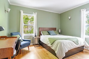 Un dormitorio con cama, escritorio, silla, ventana con cortinas y vista a la vegetación.