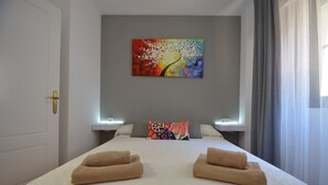 A hotel room with a bed, two towels, a colorful painting on the wall, and a window with curtains. - Alcaudón 86 - Two Bedroom Apartment, Sleeps 4 (Punta del moral)