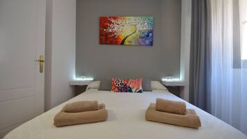Quarto de hotel com cama, duas toalhas, um quadro colorido na parede e uma janela com cortinas.