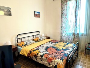 Een slaapkamer met een bed, nachtkastje, stoel, raam met gordijnen en een aan de muur hangend schilderij.