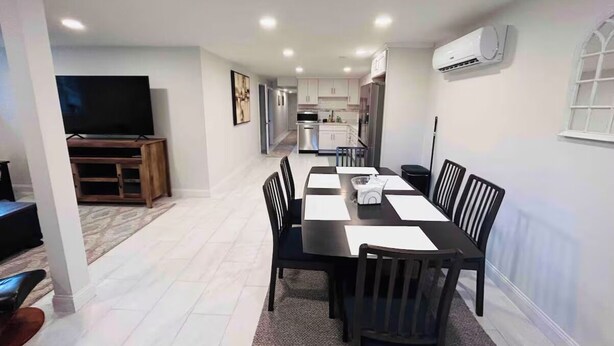 Uma área de refeições moderna com uma mesa comprida, cadeiras e uma televisão ao fundo.