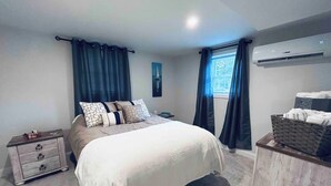 Quarto com cama, mesa de cabeceira, cômoda, janela com cortinas e ar-condicionado.