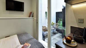 Ein modernes Zimmer mit einem Bett, einem Flachbildfernseher, einem kleinen Tisch mit Wasserkocher und Blick auf eine Terrasse mit Stühlen und Pflanzen.