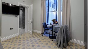 Ein Zimmer mit an der Wand befestigtem Fernseher, einem Essbereich mit Tisch und Stühlen sowie einem Fenster mit Vorhängen.