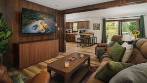 Una sala moderna con un televisor grande, paneles de madera, un sofá, una mesa de centro y vistas a la cocina y al área exterior.