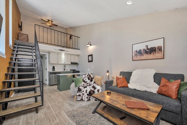 Ein Wohnzimmer mit grauem Sofa, einem Holztisch, einer Treppe und einer Küche im Hintergrund.