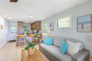 A modern living room with a sofa, a dining table with chairs, a kitchen area, and a view of the beach through the windows.
