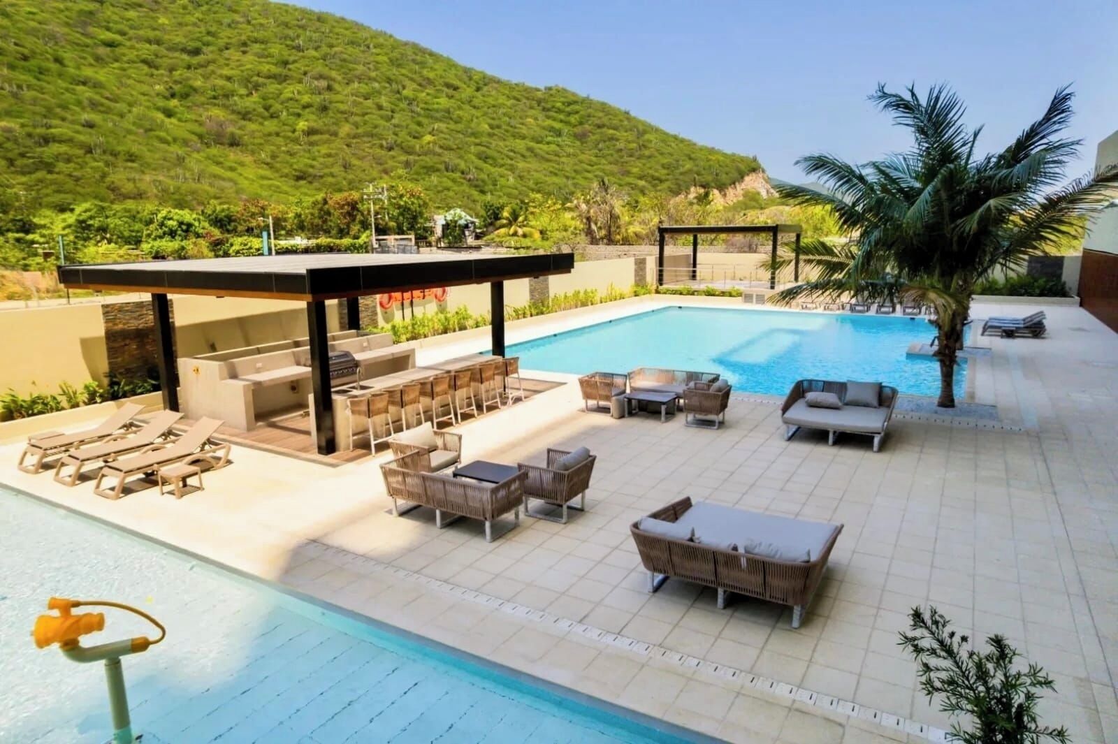 A pool area with lounge chairs and a covered seating area, surrounded by greenery and a hilly landscape.