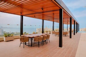 Eine Dachterrasse mit einer Holzpergola, einem langen Esstisch mit Stühlen und Blick auf Stadt und Meer.