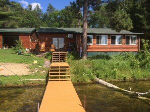 Puinen mökki, jonka edessä on laituri, joka johtaa kirkasvetiseen järveen tai mereen, ja mökkiä ympäröi vehreä luonto.