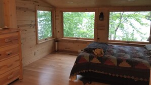 Dormitorio de cabaña de madera con cama, cómoda y ventanas amplias.