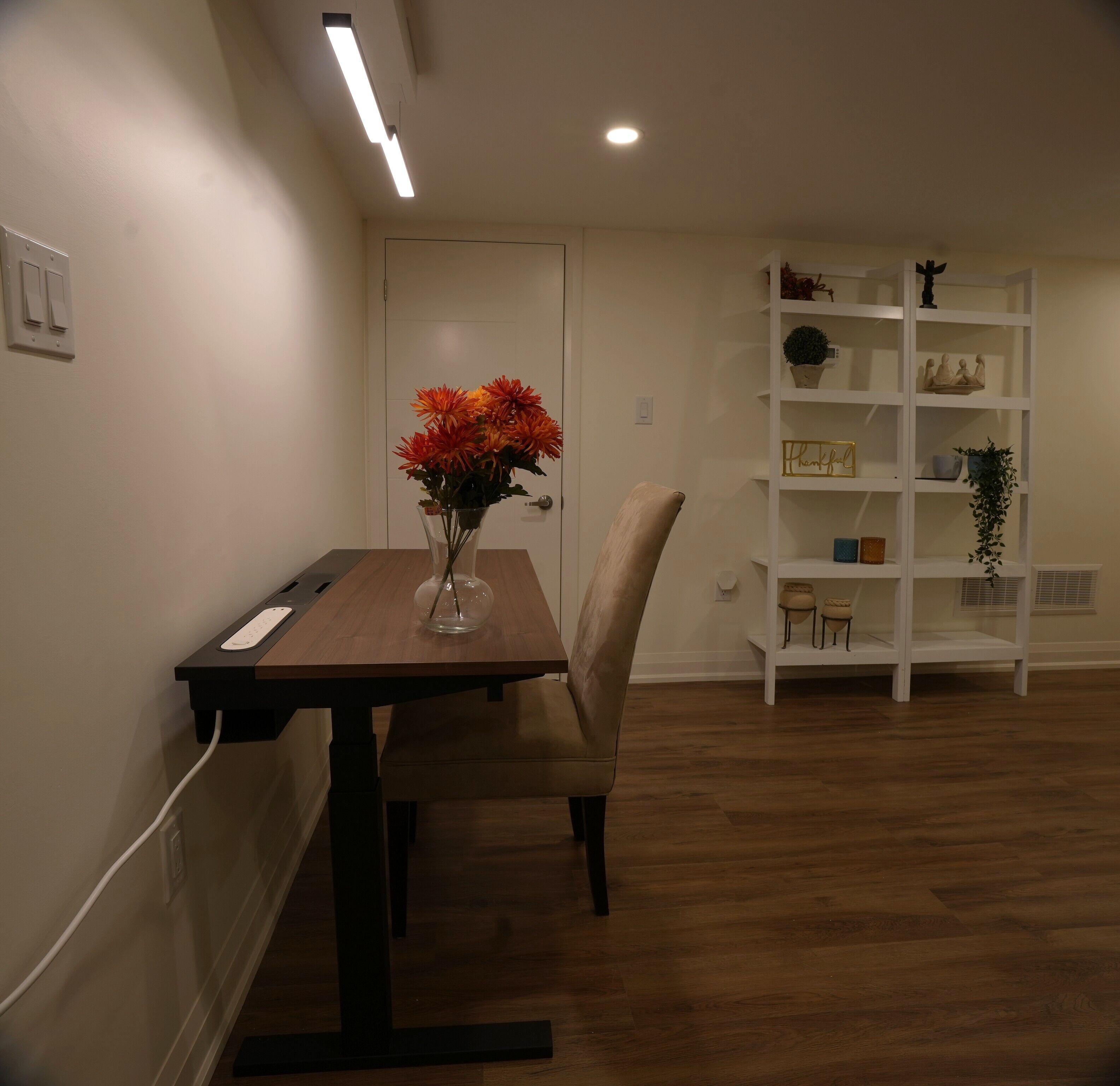 A dining area with a wooden table, a vase with flowers, a chair, a white shelf with decorative items, and a ceiling light.