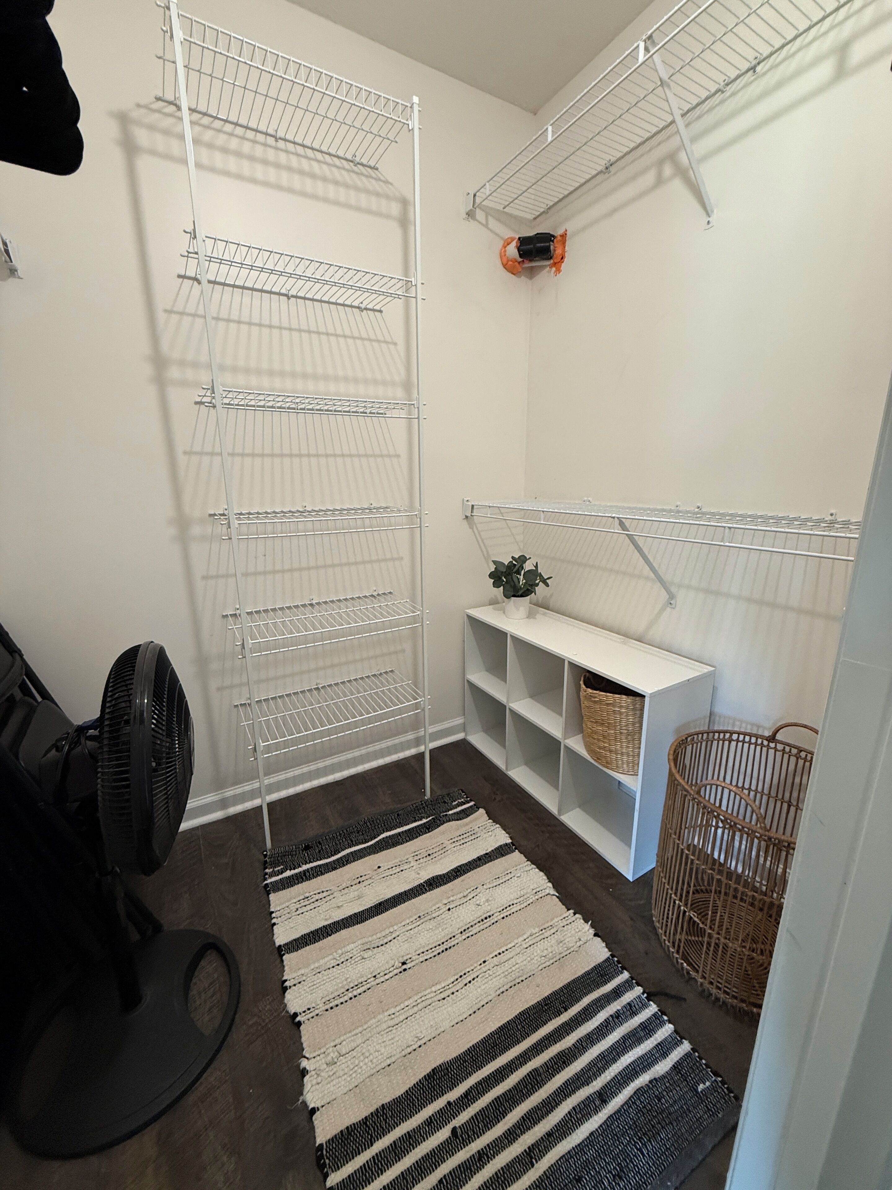 Een kleine opbergruimte met metalen schappen, een ventilator, een gevlochten mand en een gestreept vloerkleed.