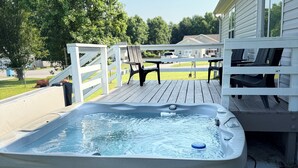 Un jacuzzi en una terraza de madera con barandilla blanca, una mesa y sillas al fondo.