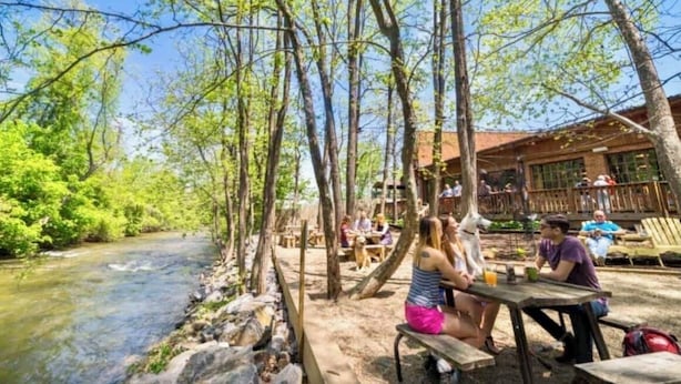A riverside outdoor seating area with wooden tables and benches, surrounded by trees and a building with large windows.