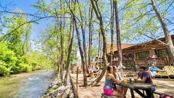 Un espace extérieur aménagé au bord de la rivière, avec des tables et des bancs en bois, entouré d’arbres et bordé d’un bâtiment doté de grandes fenêtres.