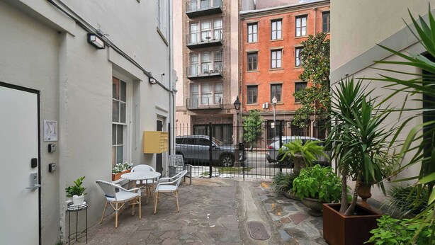 A narrow courtyard with stone flooring, a small seating area with chairs and a table, potted plants, and a building with multiple windows and balconies in the background. - Lux  Funk Suite W/2  Luxury Baths (New Orleans)
