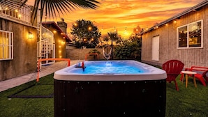Un bain à remous dans un jardin, avec vue sur le coucher de soleil, entouré d’une clôture en bois, d’un palmier et de chaises rouges.