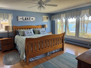 Ein Schlafzimmer mit einem hölzernen Bett, einem Nachttisch, einer Lampe, einem Fenster mit Seeblick und einem Deckenventilator.